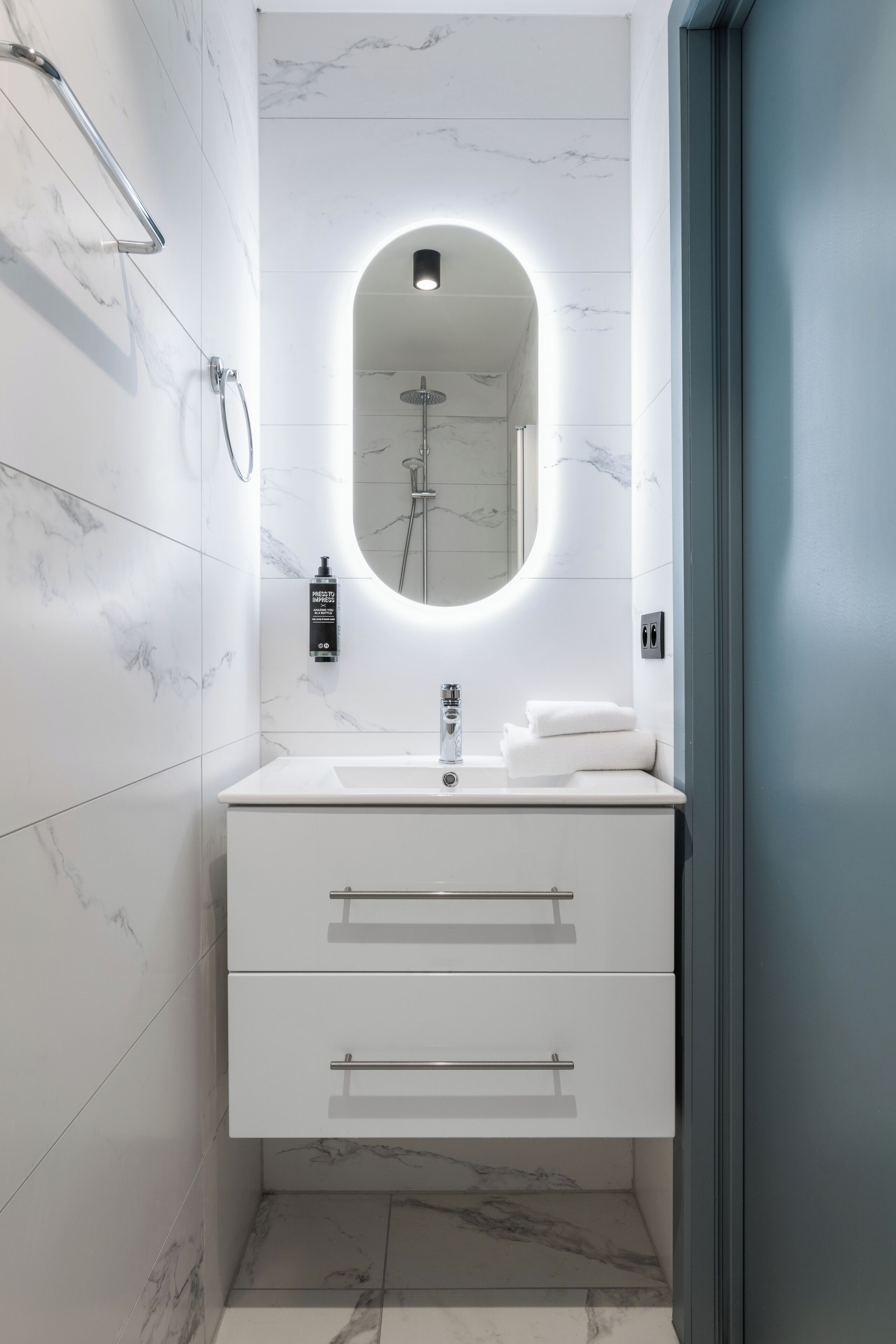 Un baño moderno con tocador blanco, espejo redondo con iluminación inferior, cabezal de ducha manual y toallero.