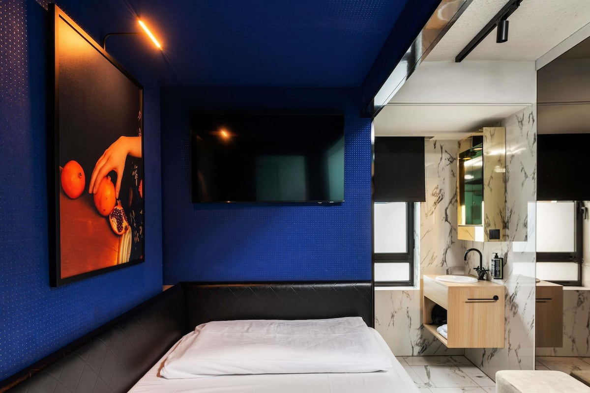 A modern hotel room with a bed, a mounted TV, a bathroom with a marble vanity, and a painting on the wall.