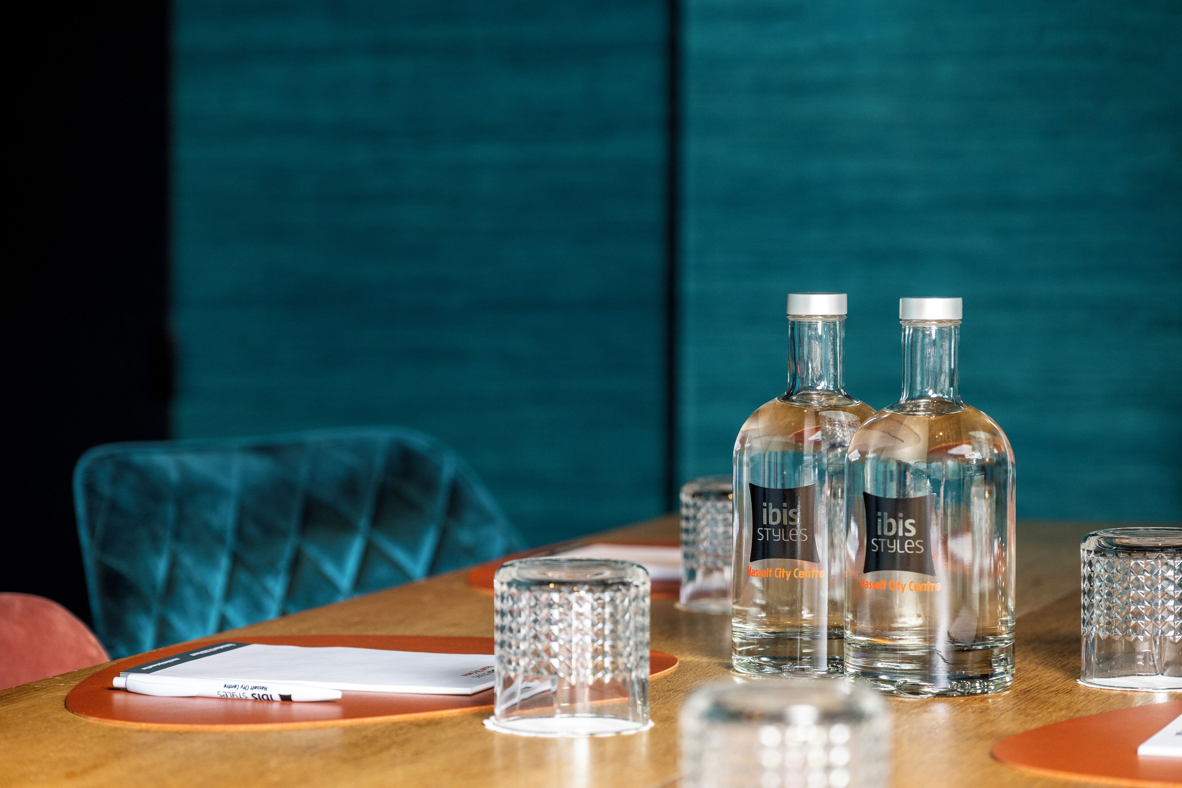 Una mesa con dos botellas de agua 'Ibis Styles', un vaso y un bolígrafo sobre una mesa de madera con fondo color aguamarina.