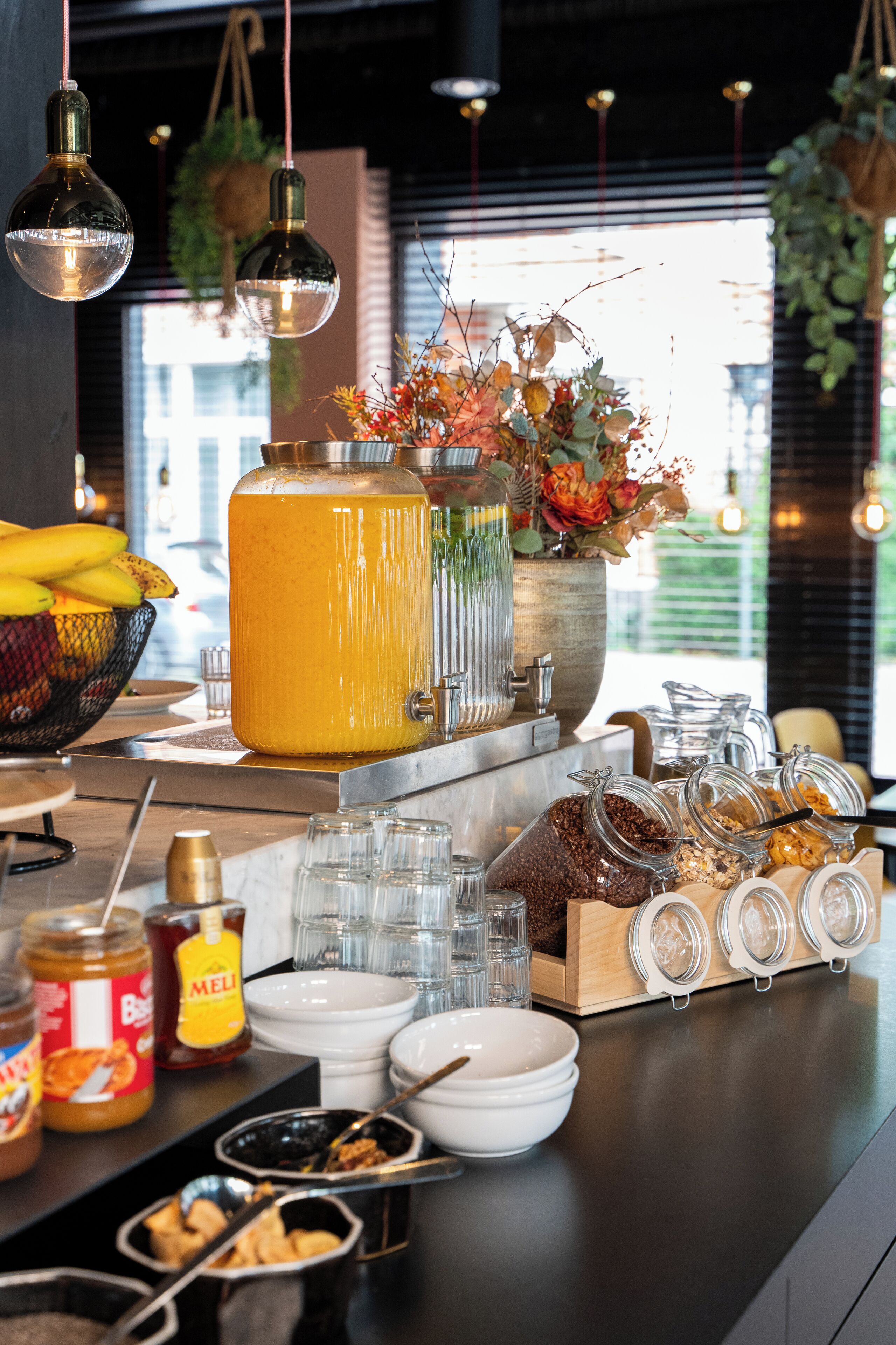 Um buffet de café da manhã com diversos alimentos, um grande dispensador de suco de laranja e luminárias penduradas.