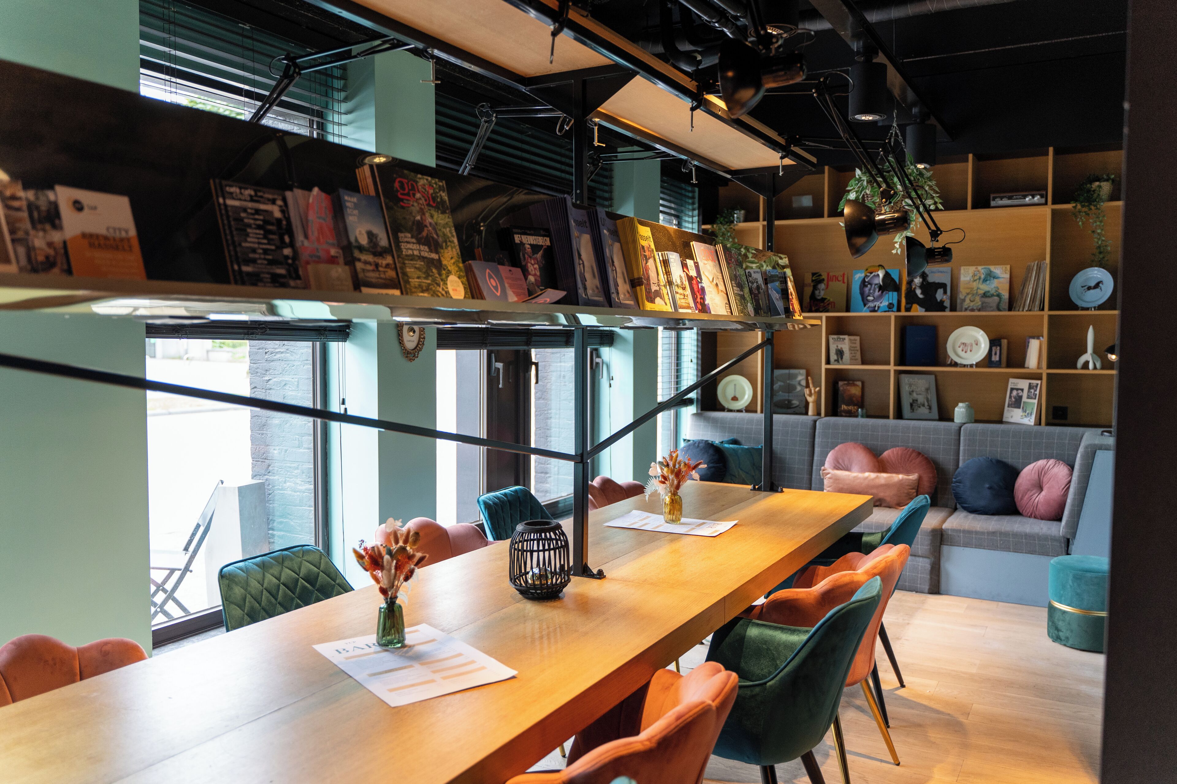 A modern interior with a long wooden table, colorful chairs, a sofa, and a bookshelf filled with books and decorative items.