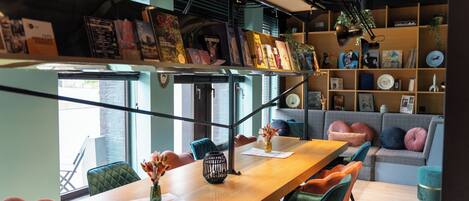 Un interior moderno con una larga mesa de madera, sillas coloridas, un sofá y una estantería llena de libros y objetos decorativos.