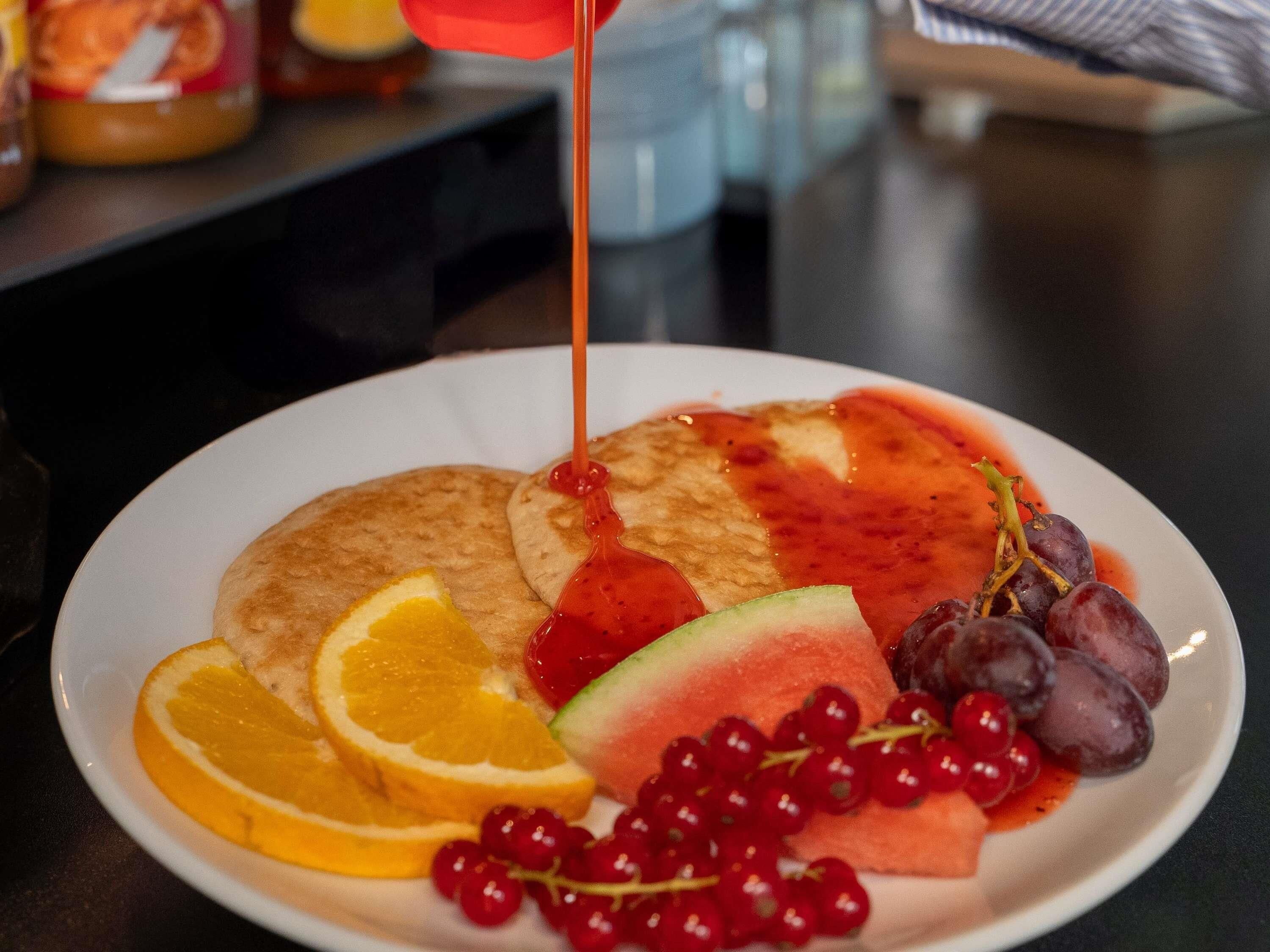 Un plato con tortitas, rodajas de naranja, sandía, uvas y un vaso de zumo de naranja.
