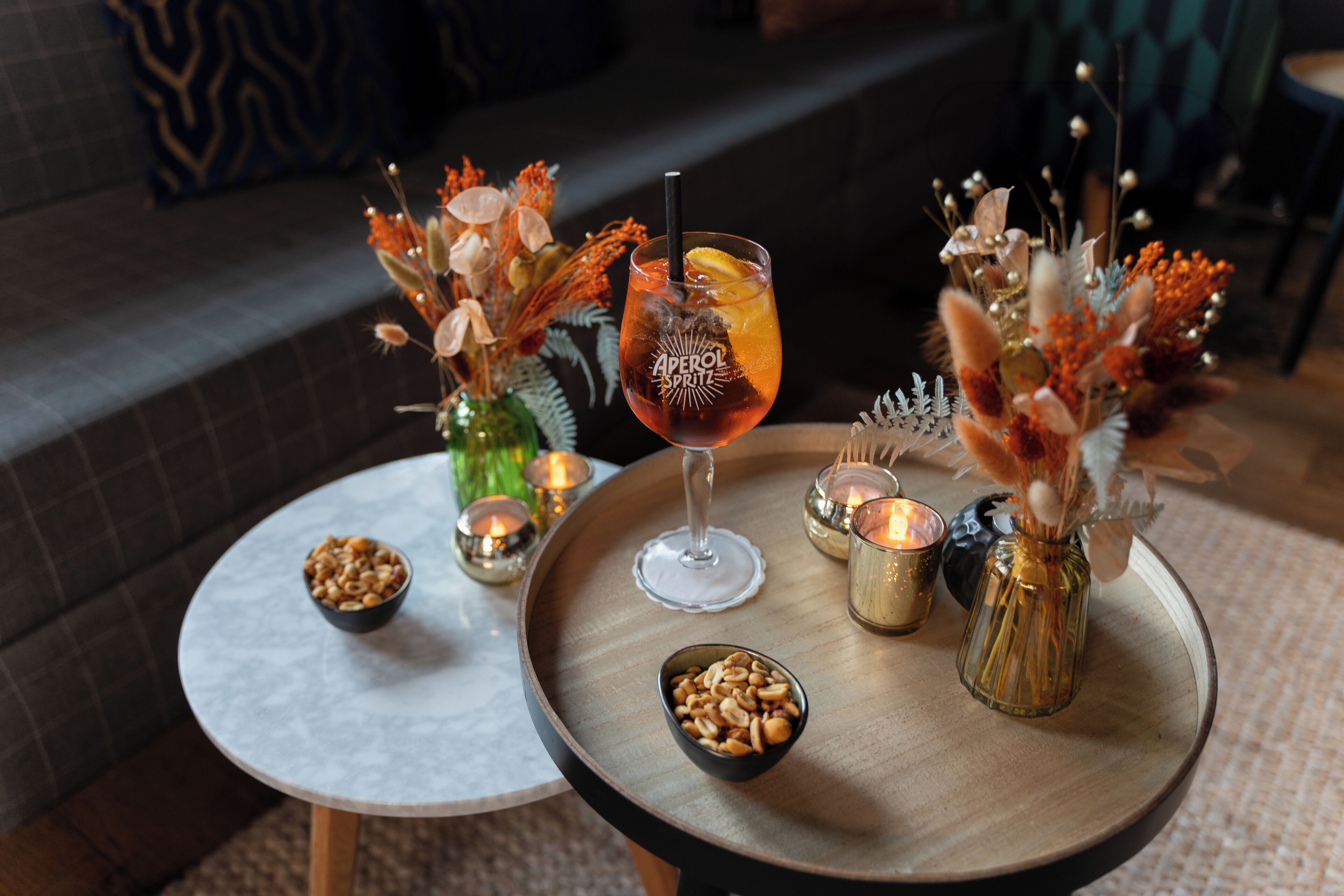 A table with a glass of Aperol Spritz, a bowl of nuts, and decorative vases with dried flowers.