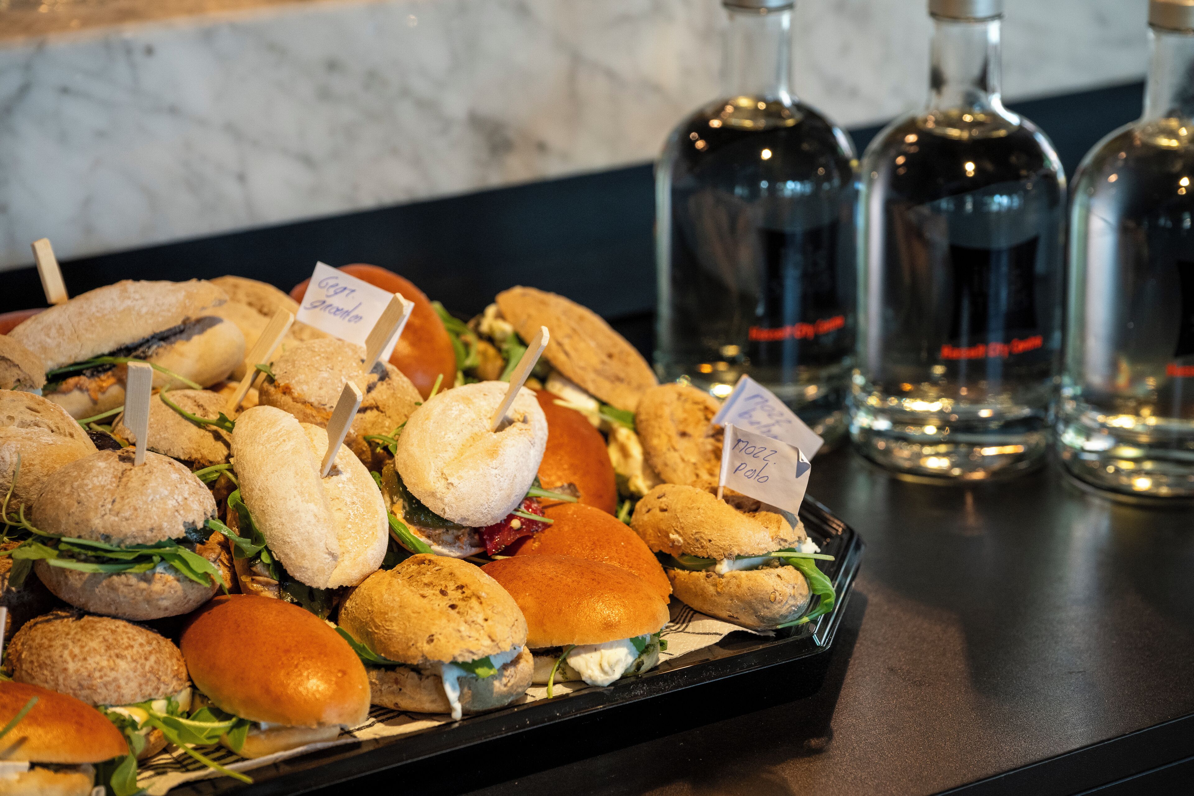 Een dienblad met sandwiches met labels, vergezeld van waterflessen op een toonbank.