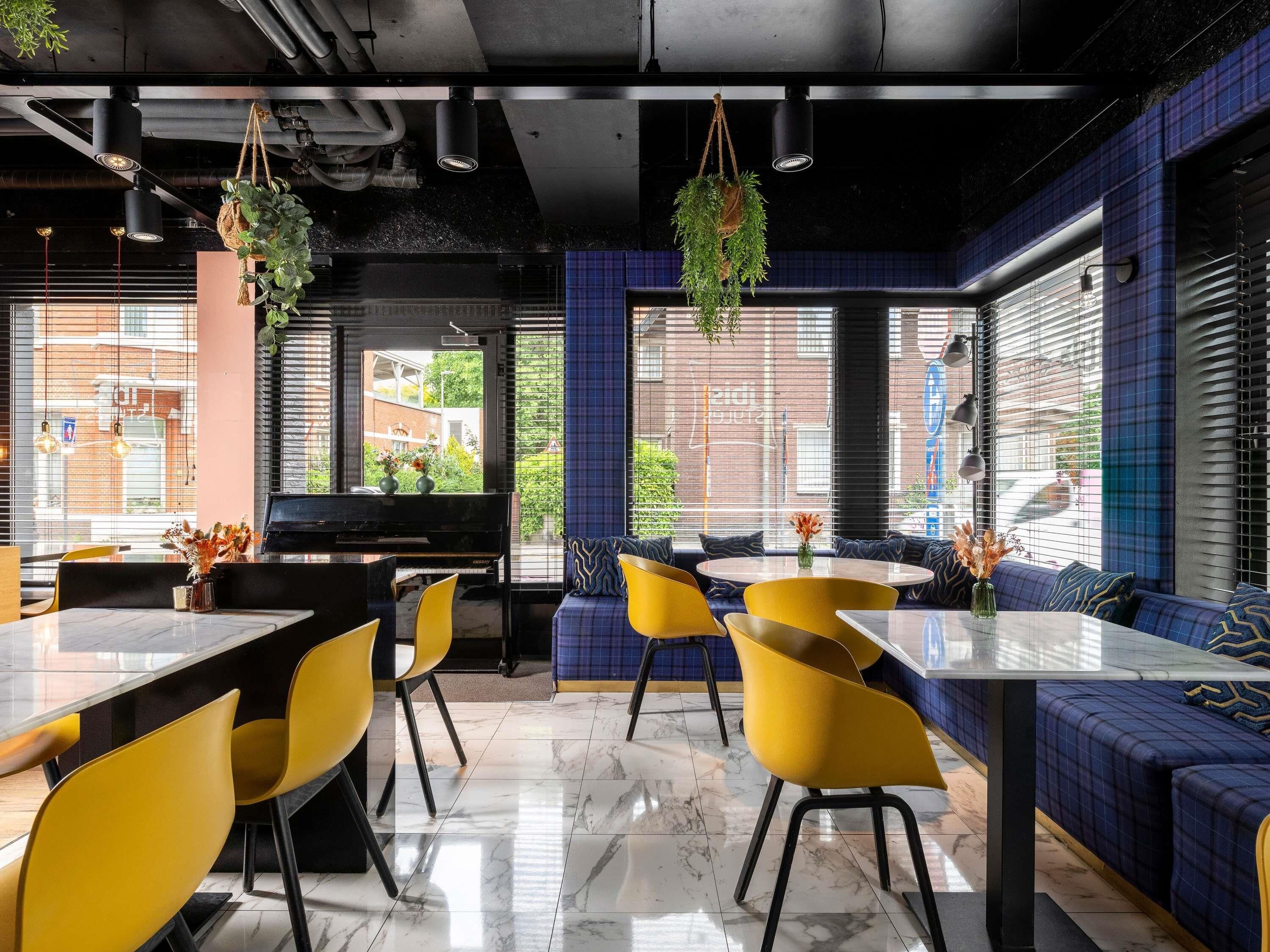 Un restaurant doté d’un sol en marbre, de chaises jaunes, de murs bleus et noirs, de plantes suspendues et offrant une vue sur la rue.