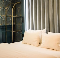 Una cama con ropa blanca y dos almohadas, un atril metálico y una cortina al fondo.