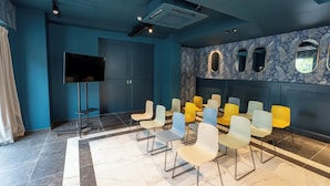 Une salle de conférence équipée d’un téléviseur à écran plat, d’un projecteur fixé au plafond et d’une rangée de chaises colorées.