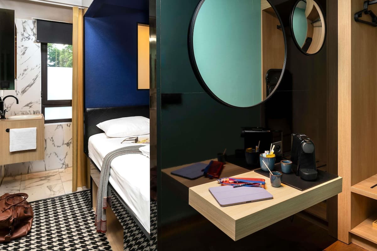A modern hotel room with a bed, a round mirror, a wooden desk with a laptop, and a bathroom with a marble sink.