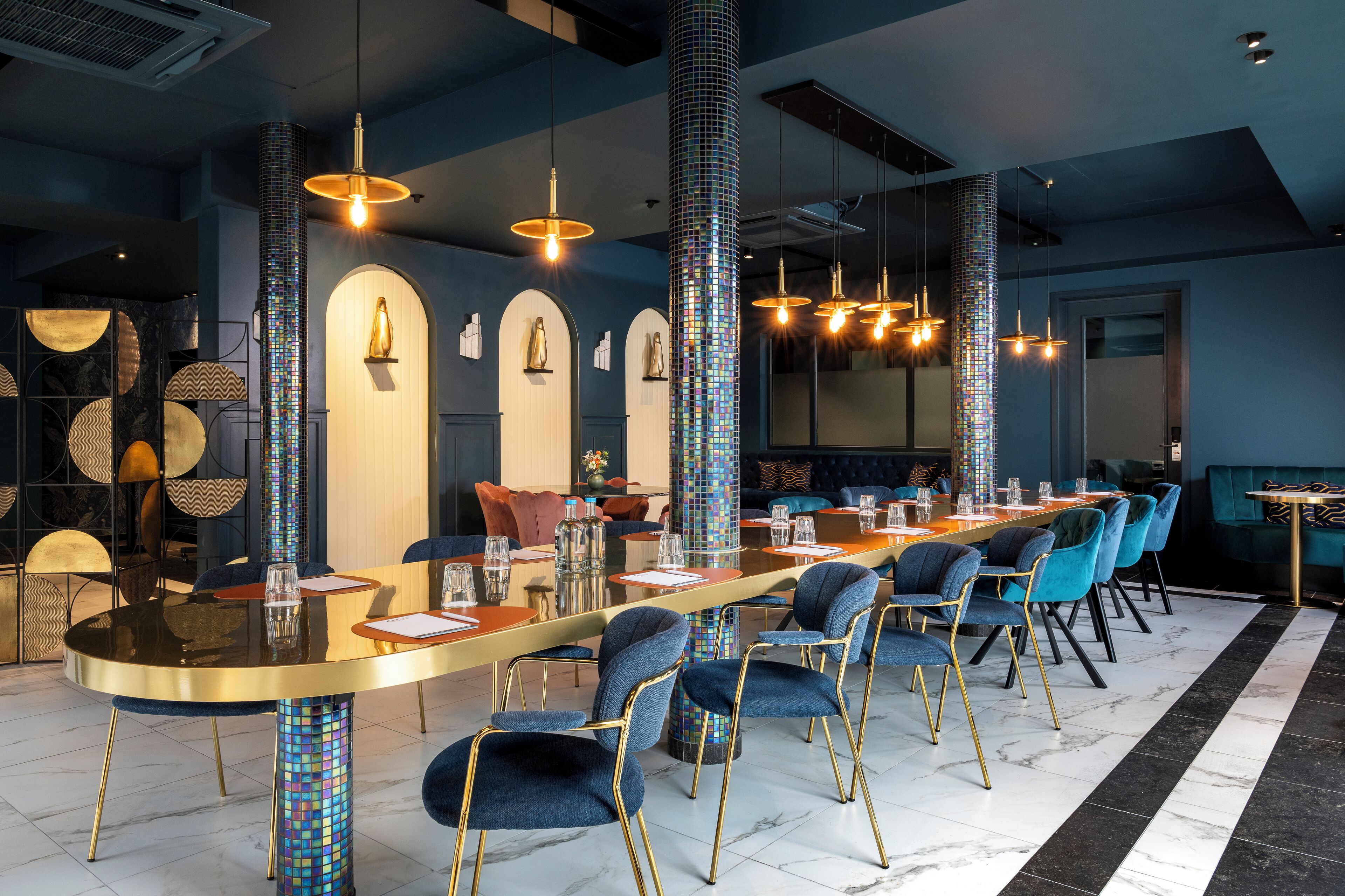 A modern dining area with a long table, blue upholstered chairs, and a mosaic column.