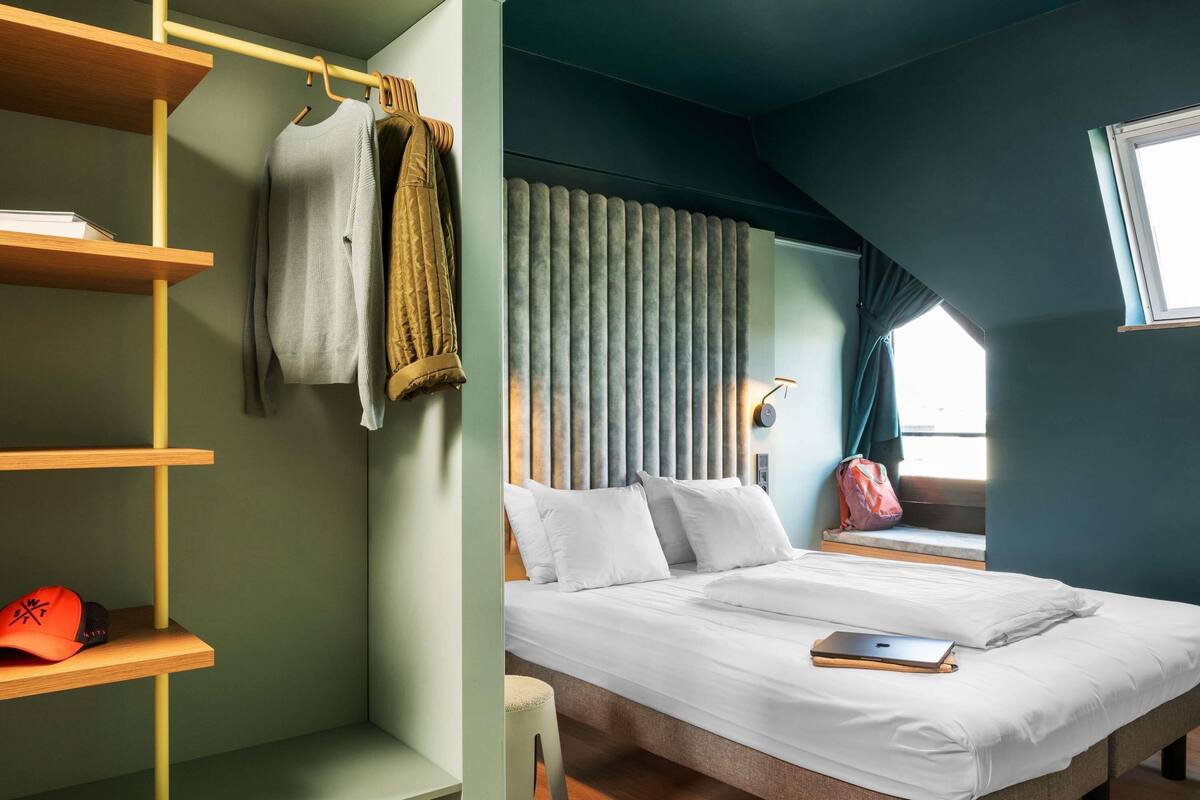 A bedroom with a bed, a shelf with clothes, a window, and a curtain.