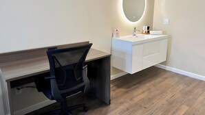 A modern bathroom with a wooden vanity, a round mirror with underglow lighting, a black office chair, and a wooden floor. - The Dixie Hollywood (Los Angeles)