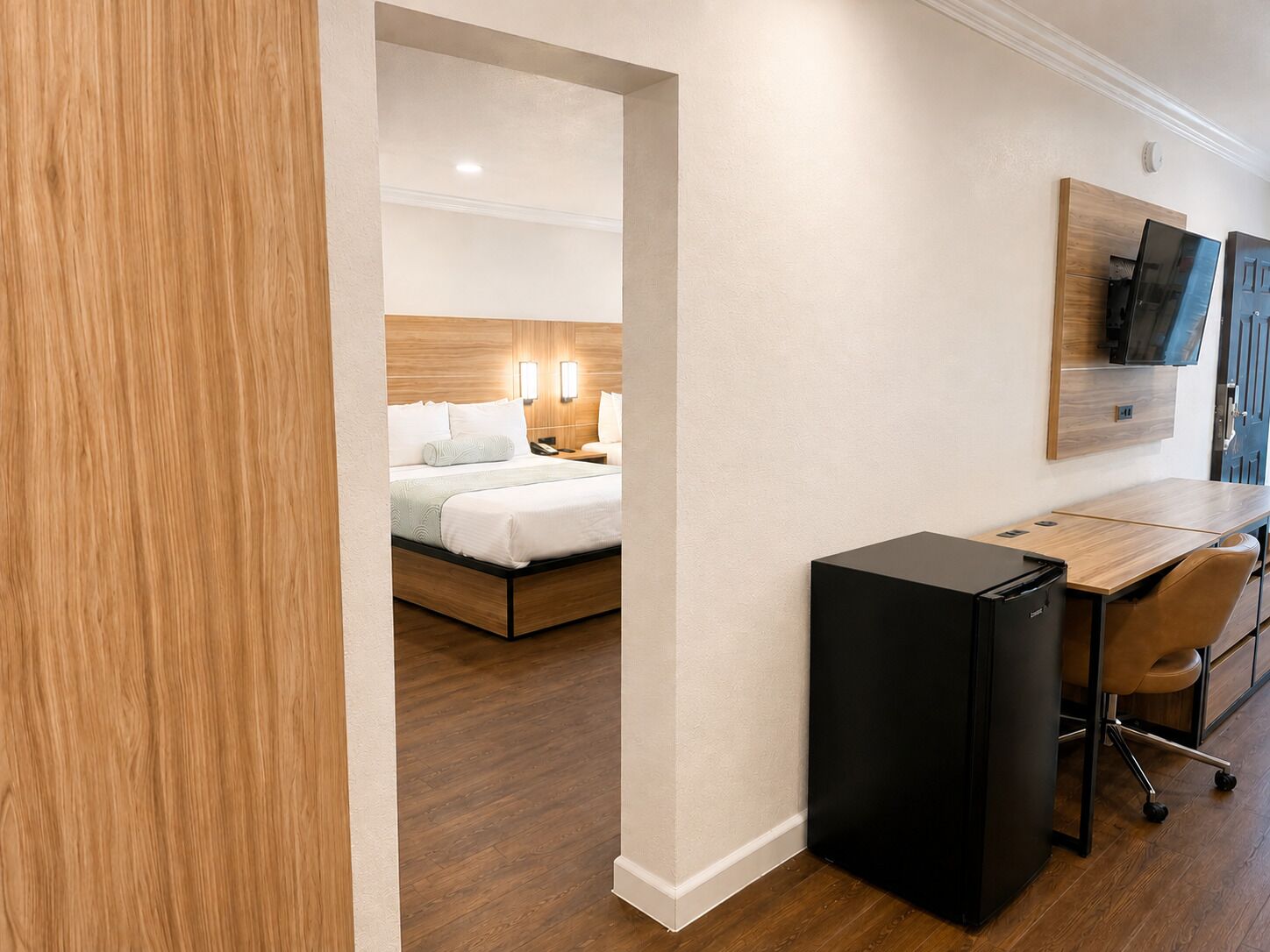 Habitación de hotel con cama, lámparas de noche, escritorio con silla, refrigerador negro y puerta de madera.
