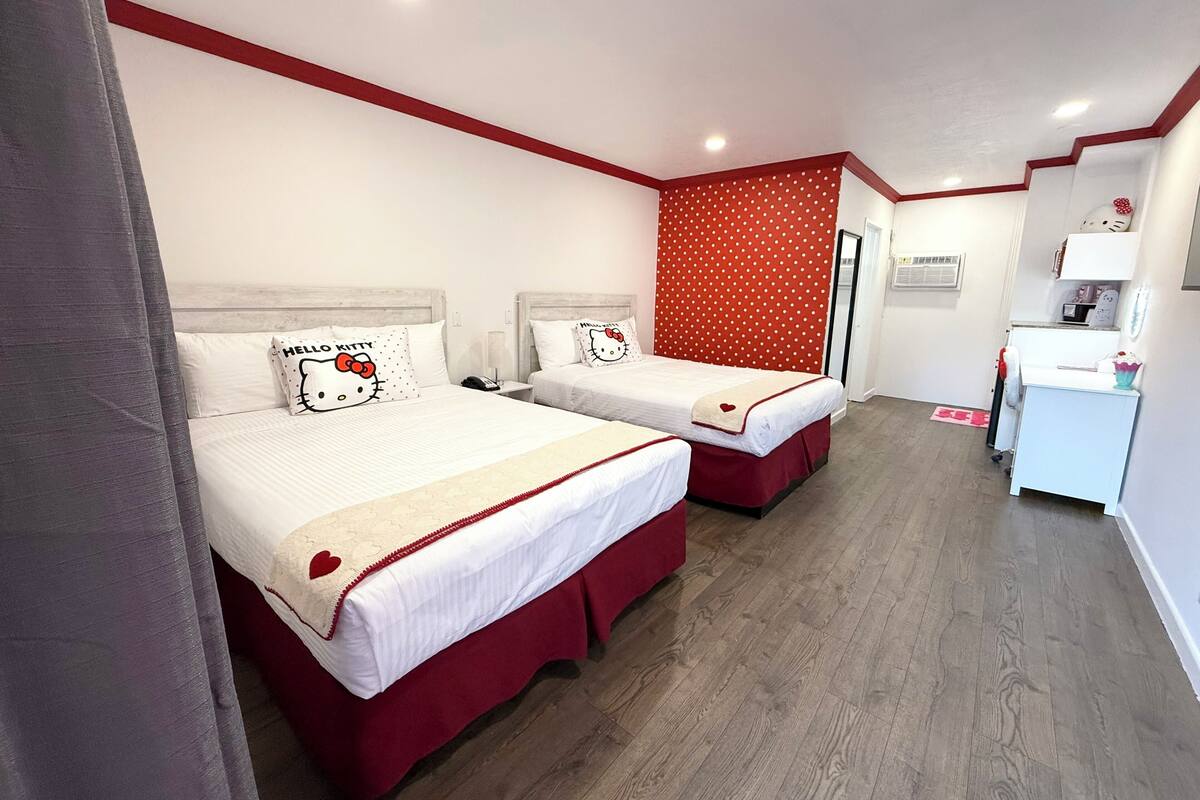 Ett hotellrum med två sängar, var och en med en Hello Kitty-kudde, en röd vägg med prickmönster och ett litet skrivbord med en lampa.