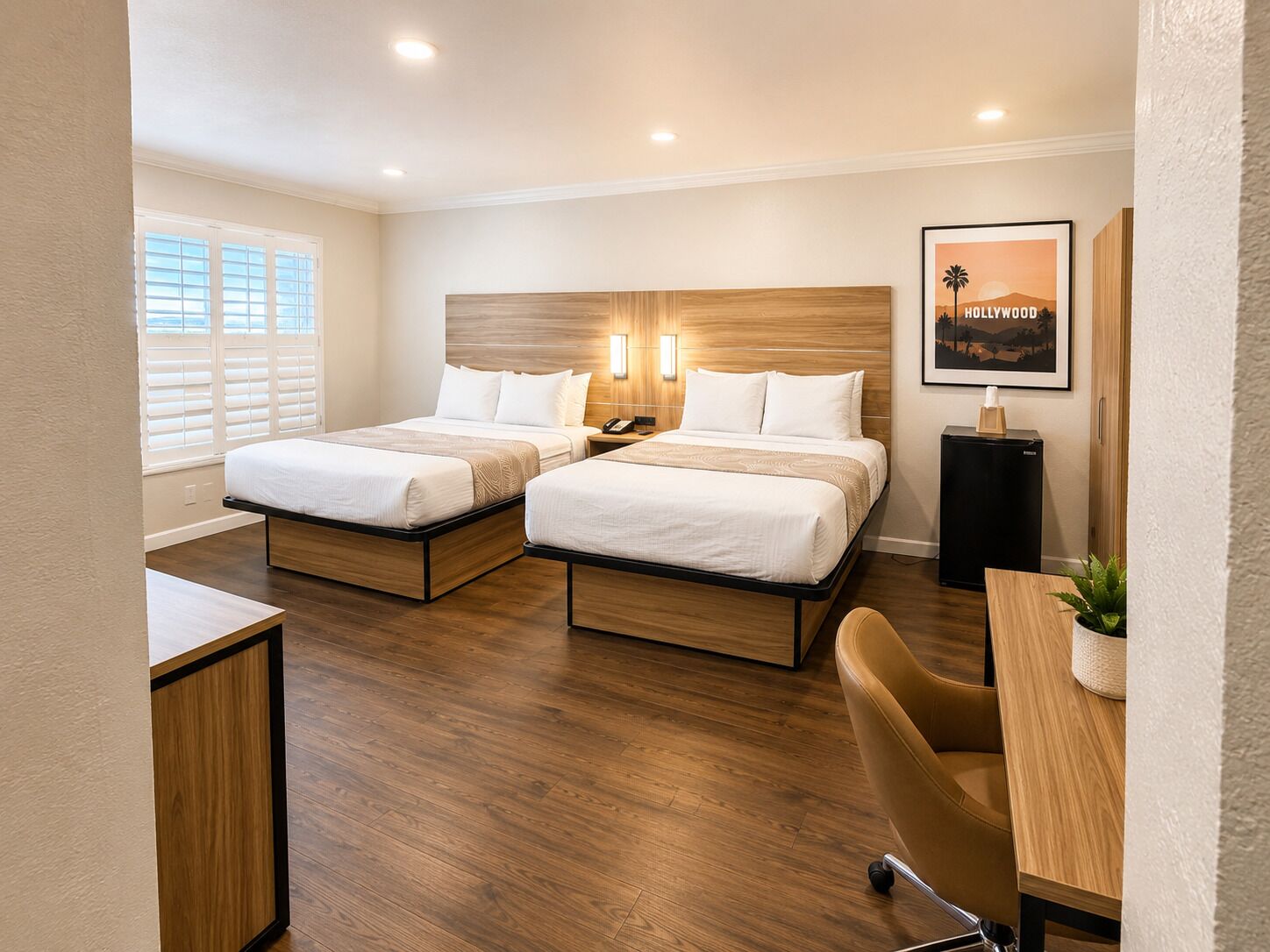 Quarto de hotel com duas camas, uma escrivaninha, uma cadeira, um pequeno frigorífico e um quadro na parede.