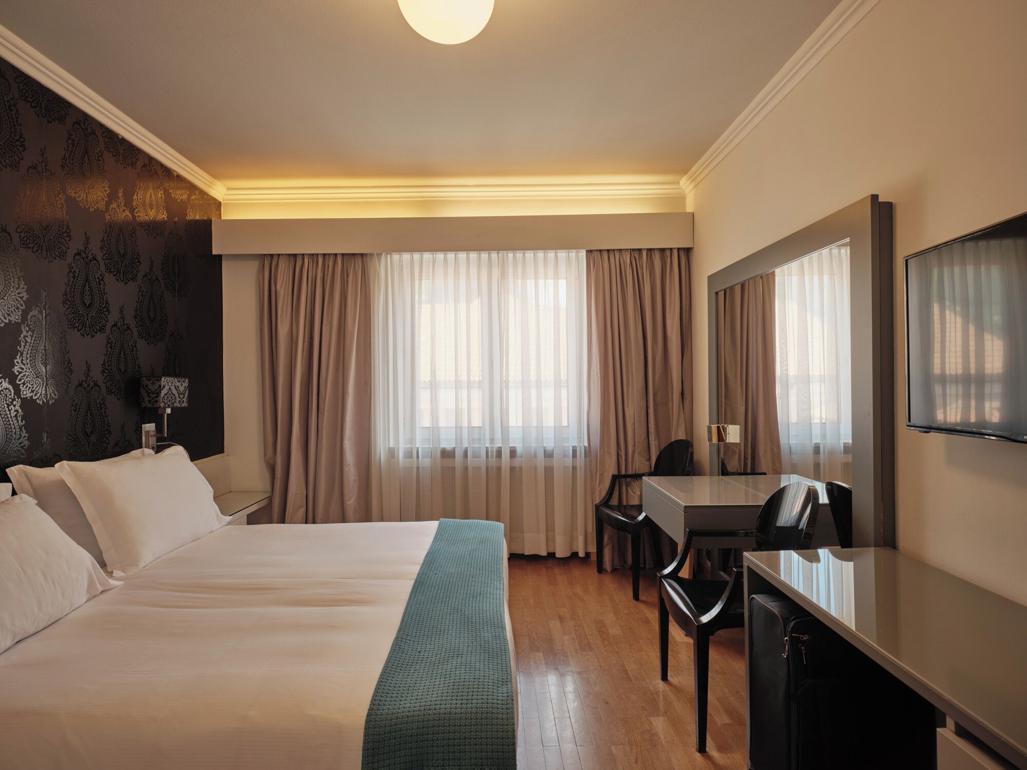 Una camera d'albergo con un letto grande, una scrivania, una sedia, una televisione e una finestra con tende.