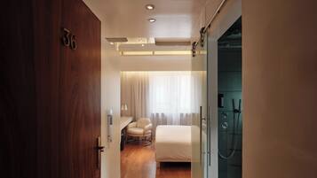 Habitación de hotel con puerta de madera, cama, silla, escritorio y zona de ducha.