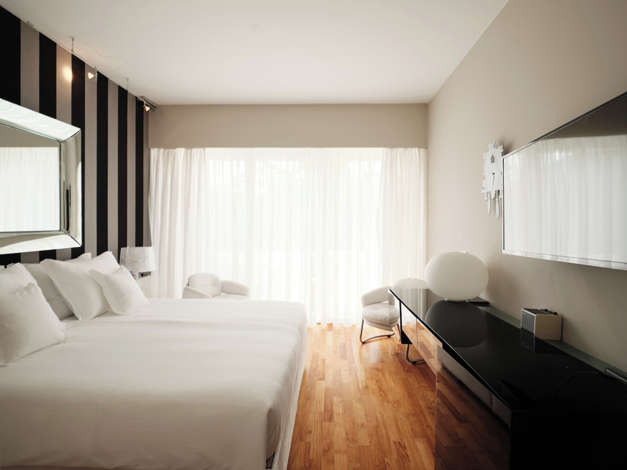Una habitación de hotel moderna con una cama grande, un escritorio, una silla, un televisor y un amplio ventanal con cortinas.