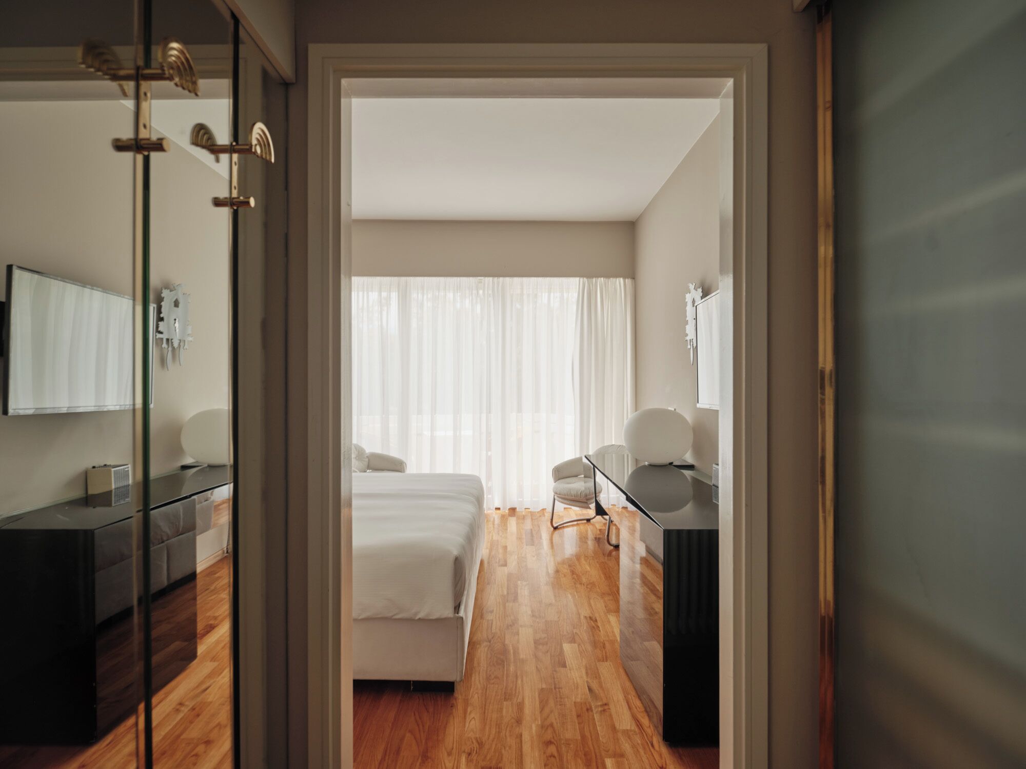 A modern hotel room with a bed, a desk, a chair, and a large window with sheer curtains.
