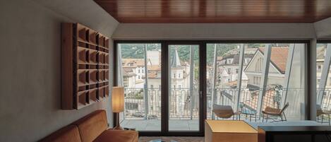 A modern living room with a brown sofa, a glass coffee table, a wooden shelf, and a view of a cityscape through large windows.