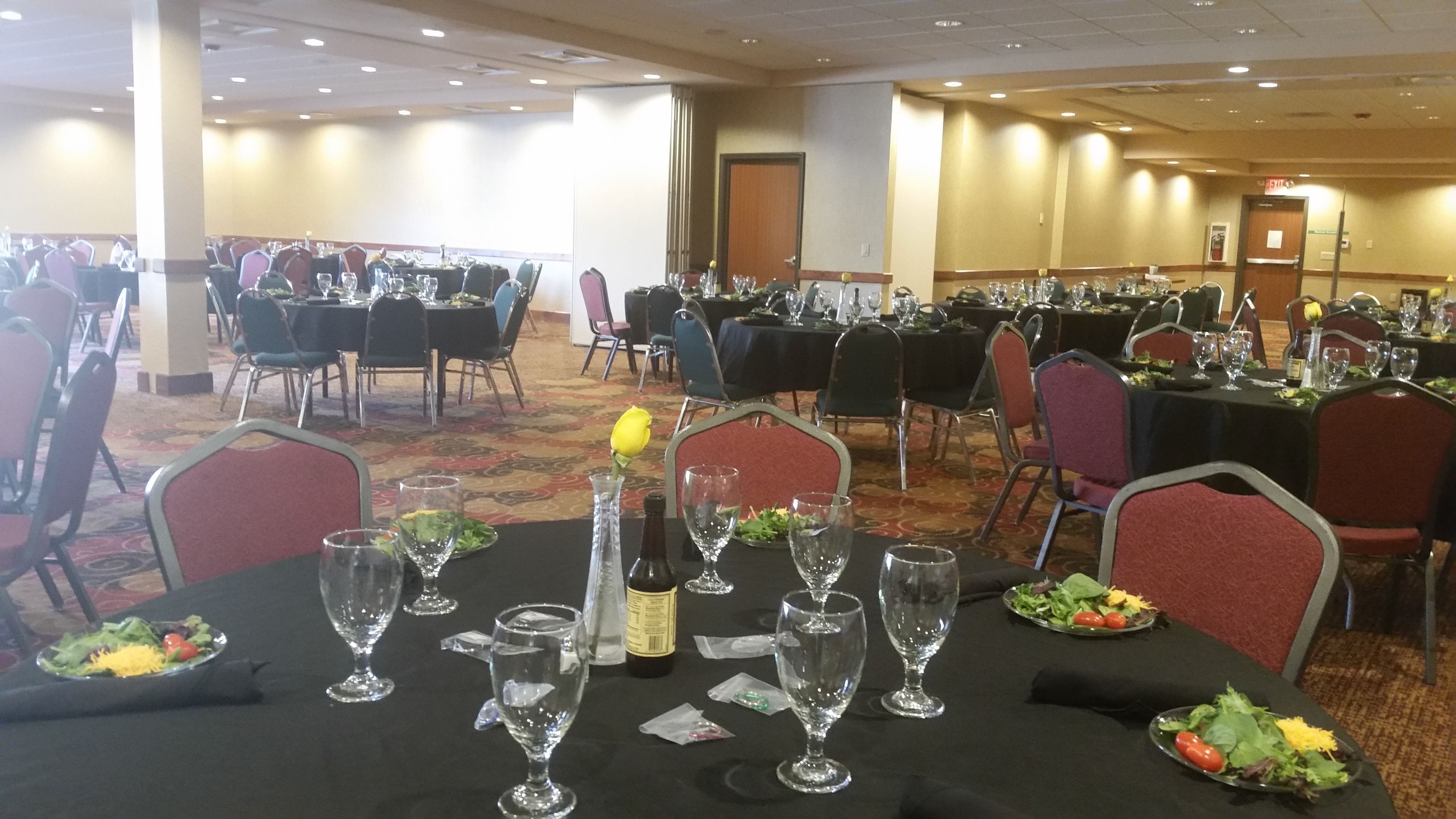 A banquet hall with multiple tables set for dining, each with a bottle of wine, glasses, and plates of salad.