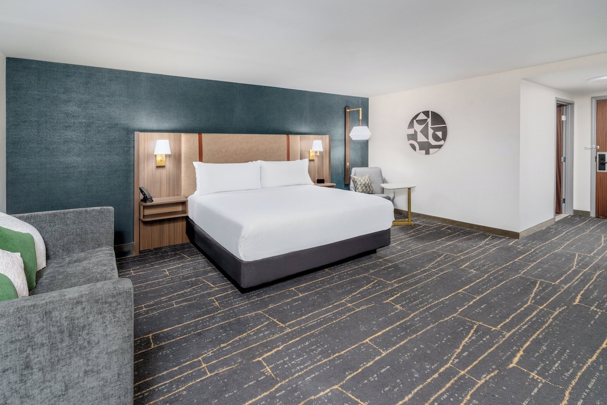 A hotel room with a large bed, a desk with a lamp, a chair, and a wall-mounted artwork.
