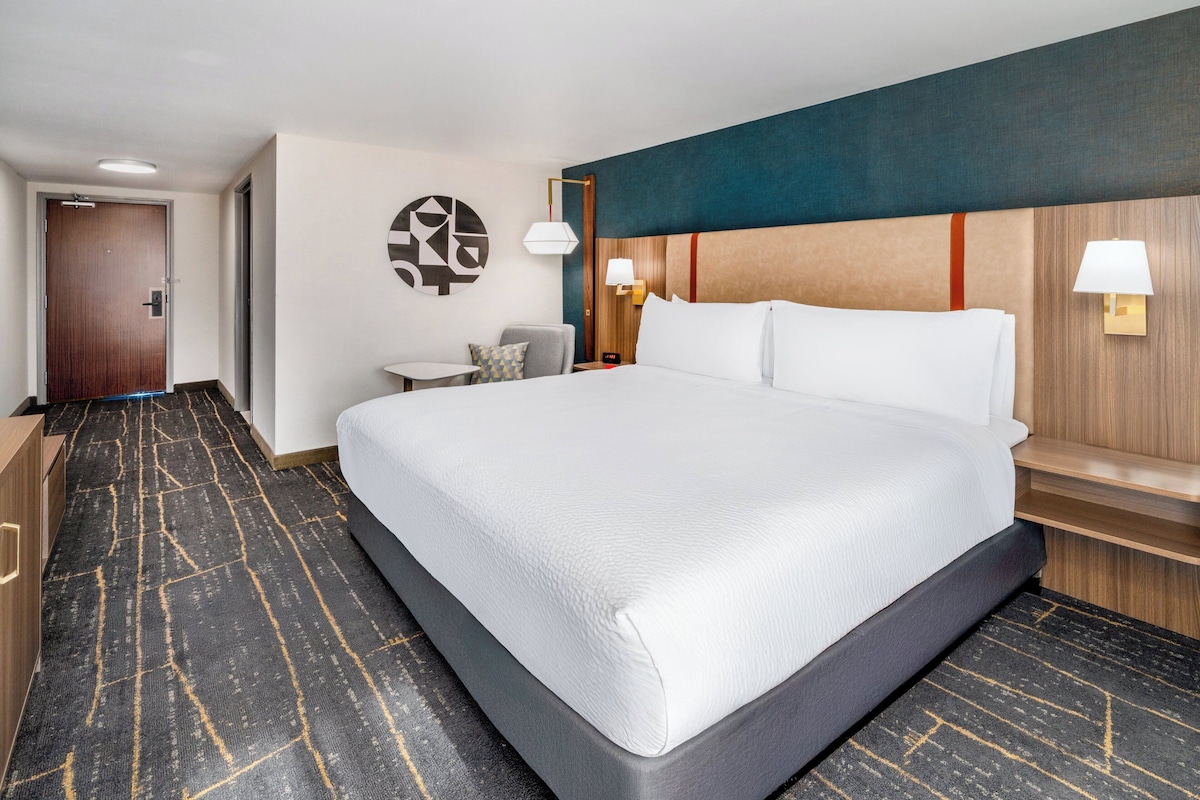 A hotel room with a large bed, a desk, a chair, and a wall-mounted lamp.