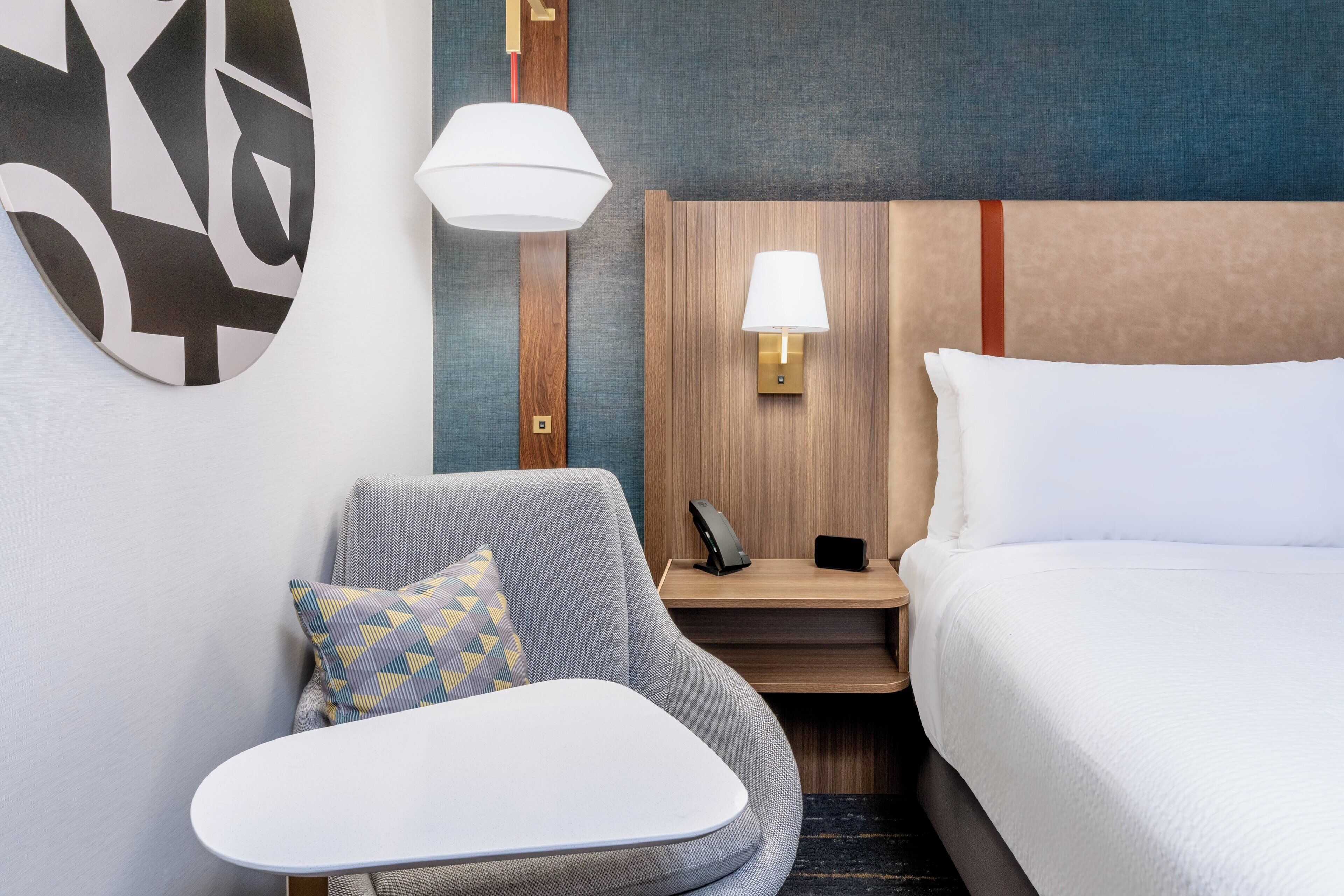 Une chambre d’hôtel avec un lit, une chaise, une petite table, une lampe et un téléphone posé sur une table de chevet en bois.