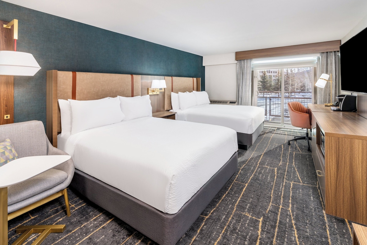 A hotel room with two beds, a desk, a chair, and a TV.