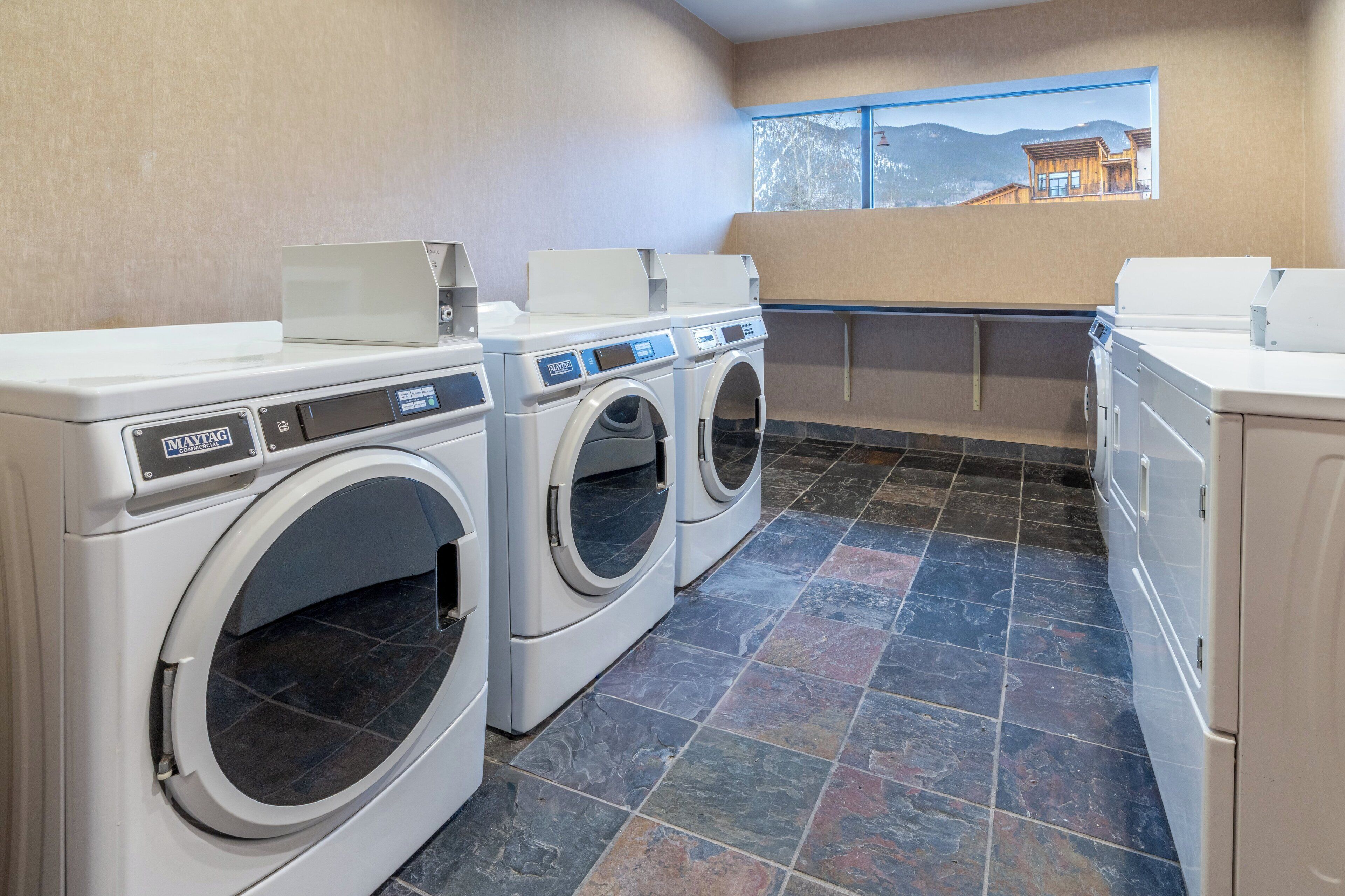 A laundry room with two front-loading washers and dryers, a window with a view of a mountain cabin, and a tiled floor.