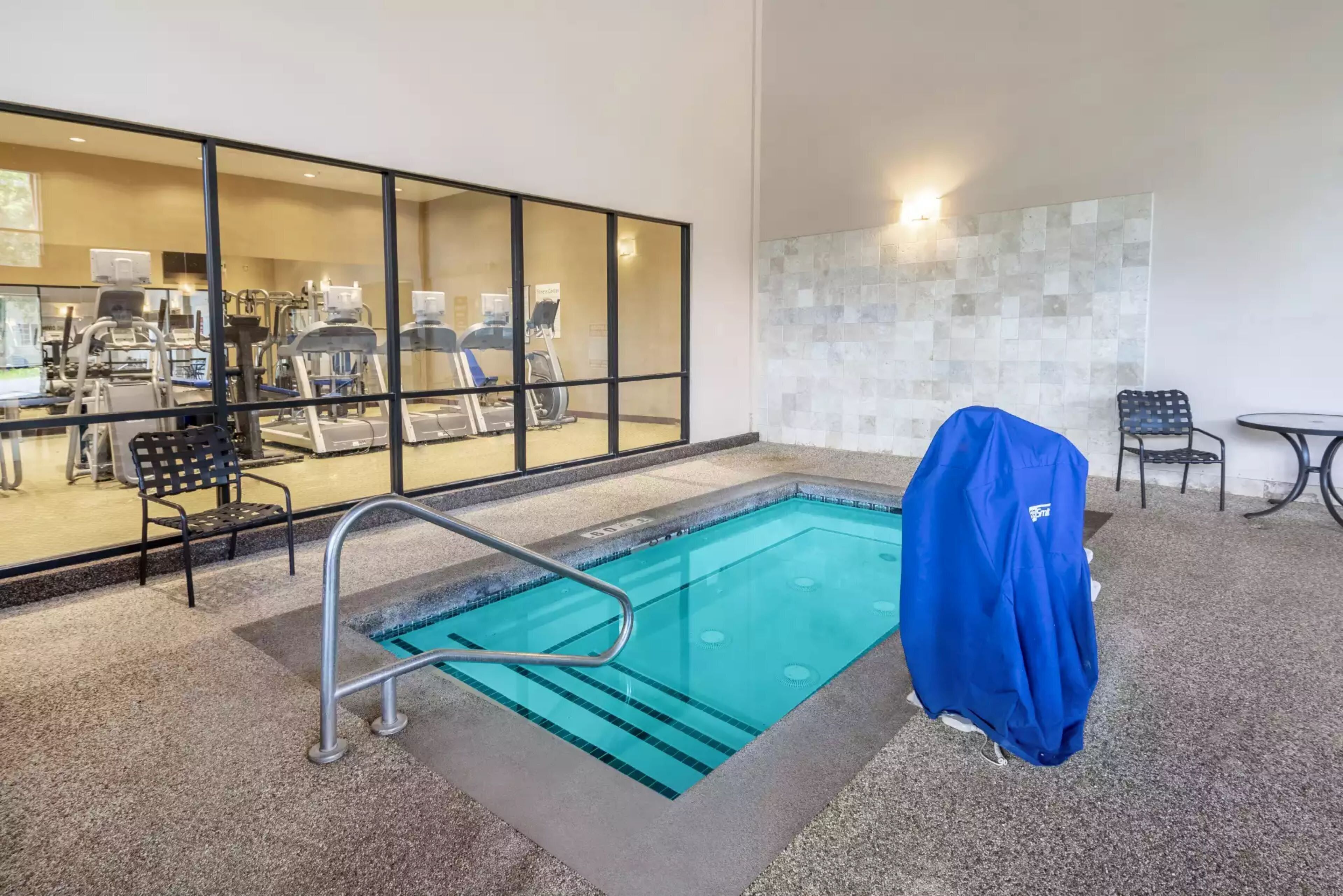 An indoor swimming pool with a covered bench, a chair, and exercise equipment in the background.