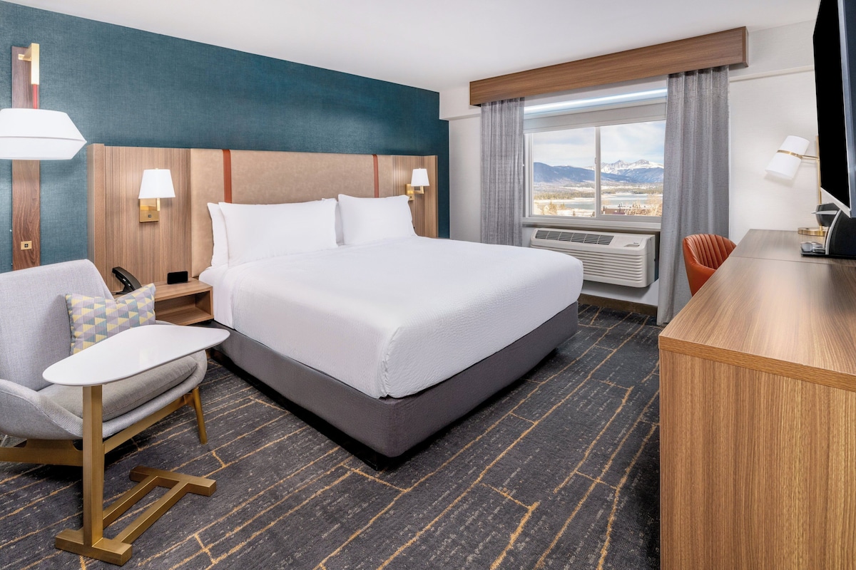 A hotel room with a large bed, a desk, a chair, and a view of mountains through the window.