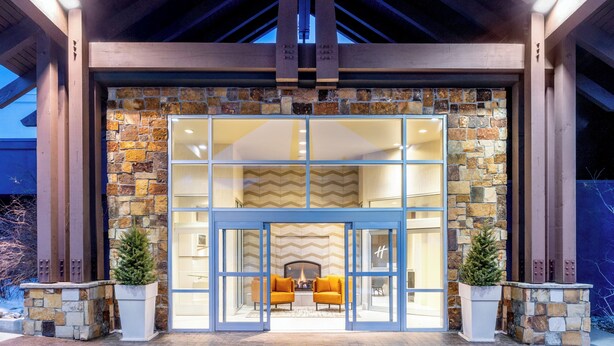 A stone facade with a glass entrance, two potted plants, and a fireplace with two chairs. - Holiday Inn Frisco by IHG (Frisco)