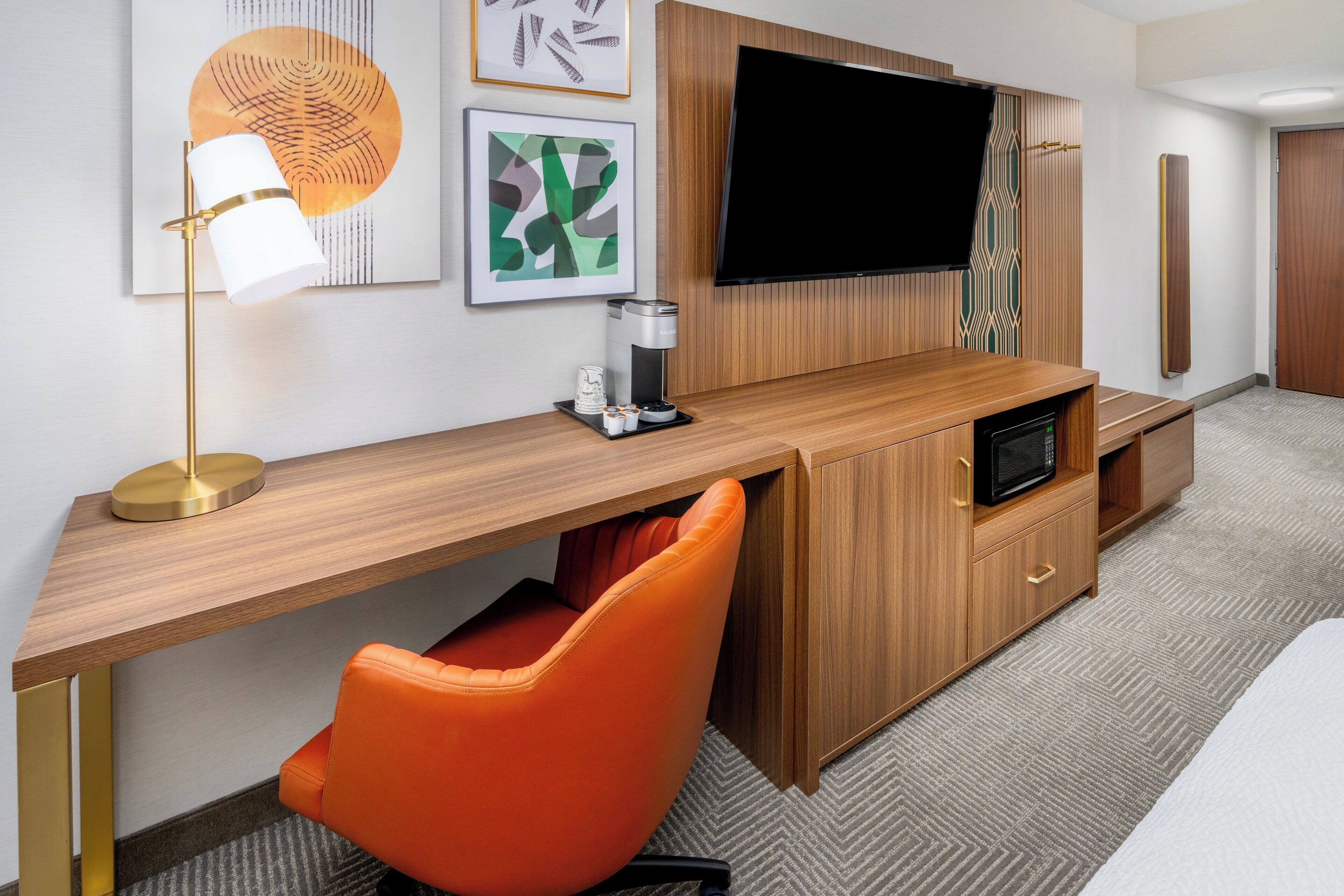 A hotel room with a wooden desk, orange chair, flat-screen TV, coffee maker, and a microwave.