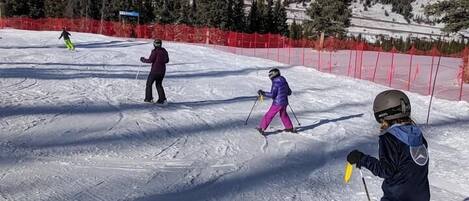 Une piste de ski avec des skieurs, une barrière de sécurité rouge et des montagnes enneigées en arrière-plan.