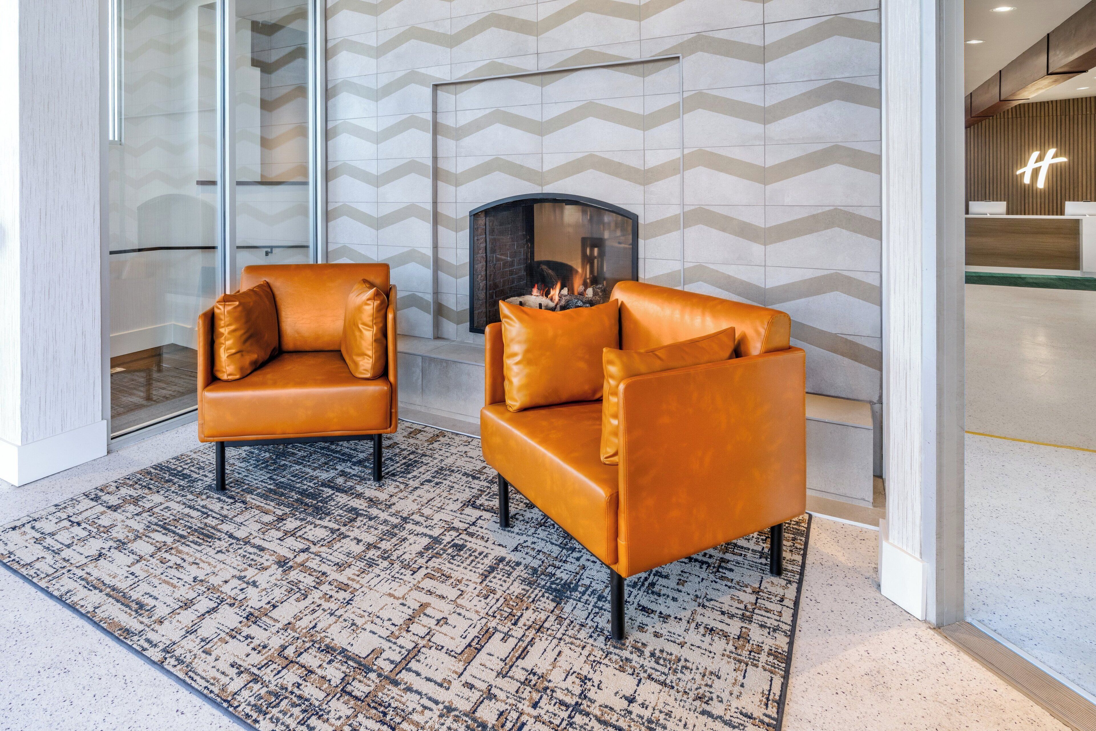 A modern lobby with two leather armchairs facing a fireplace, a patterned rug, and a chevron-patterned wall.
