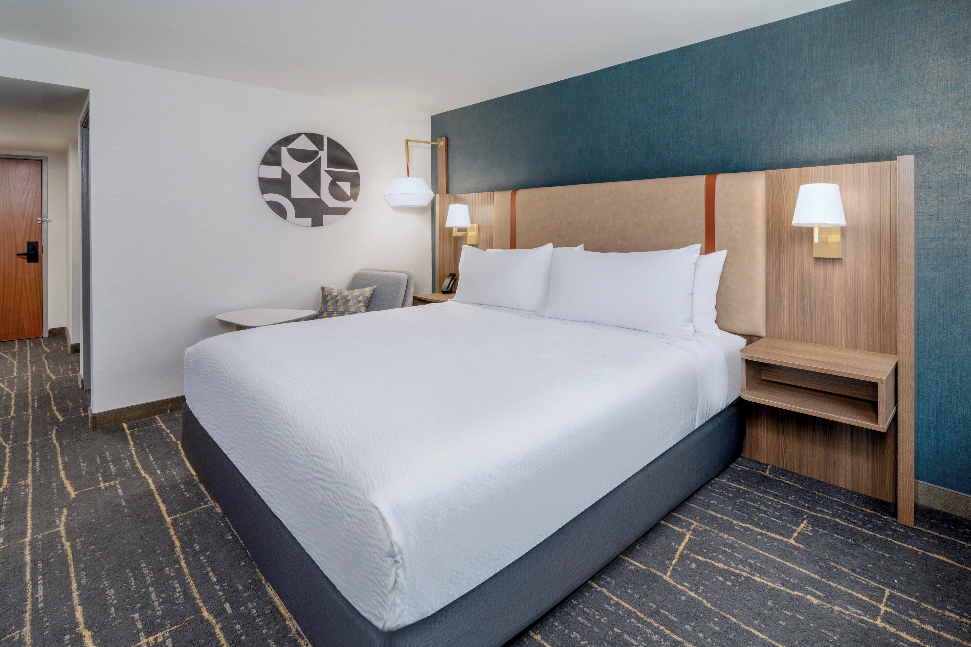 A hotel room with a large bed, a nightstand, a chair, and a wall-mounted lamp.