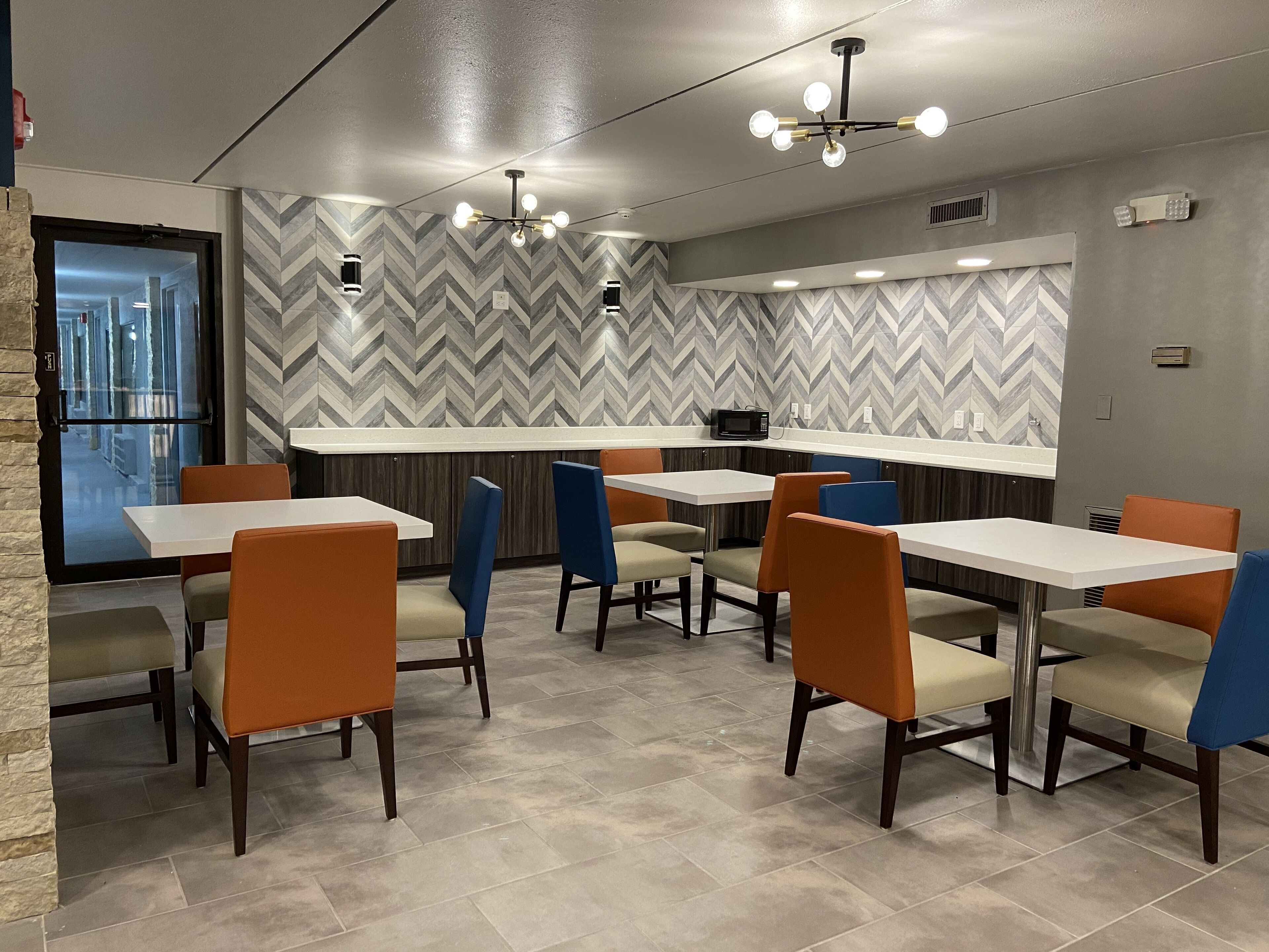 A modern restaurant with a herringbone patterned wall, white tables, and orange and blue chairs.