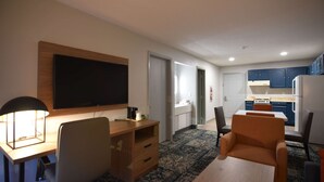 Une chambre d’hôtel moderne dotée d’un bureau en bois, d’un téléviseur à écran plat, d’un coin repas avec une table et des chaises, et d’un coin cuisine avec des placards et des appareils électroménagers.