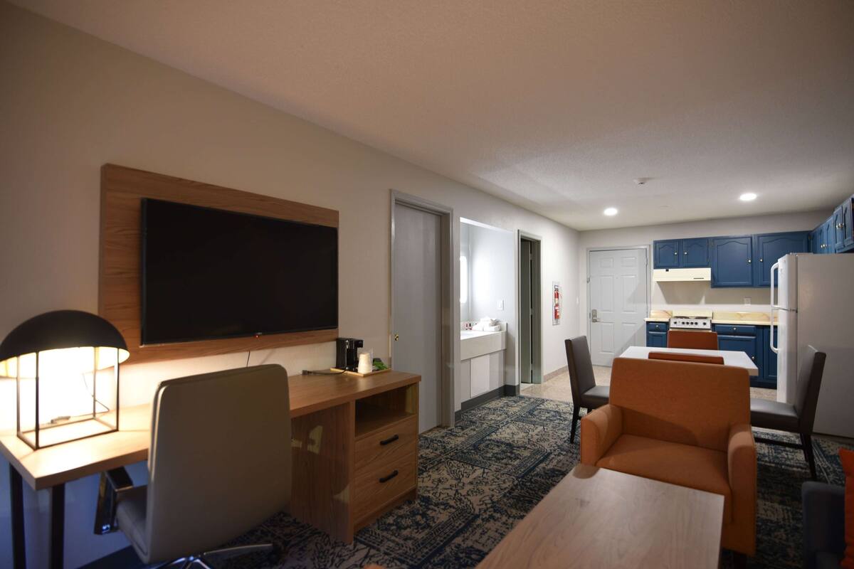 Een moderne hotelkamer met een houten bureau, een flatscreen televisie, een zithoek met een tafel en stoelen, en een keuken met kastjes en apparatuur.