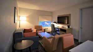 Une chambre d’hôtel compacte comprenant un canapé, des fauteuils, une petite table, un bureau avec un ordinateur, une télévision et une fenêtre avec des stores.