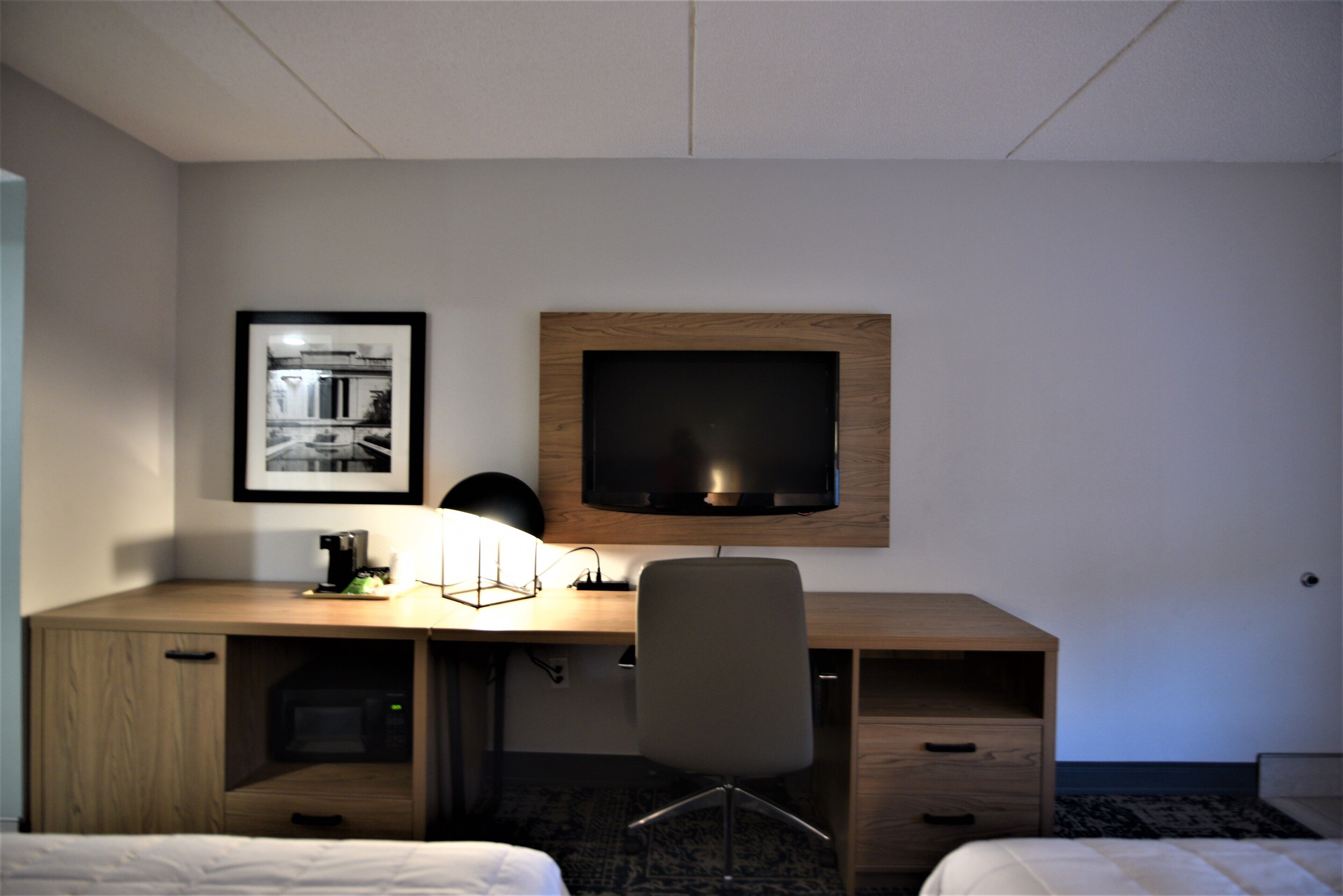 A hotel room with a wooden desk, a flat-screen TV, a framed photo on the wall, a lamp, and a chair.