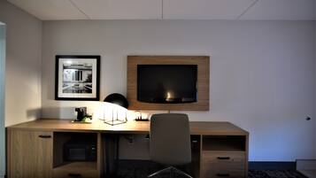 A hotel room with a wooden desk, a flat-screen TV, a framed photo on the wall, a lamp, and a chair.