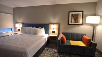 Una habitación de hotel con una cama grande, dos mesitas de noche con lámparas, un sofá gris con un cojín naranja, un cuadro en la pared y una alfombra estampada.