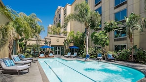 A swimming pool with lounge chairs and umbrellas, surrounded by palm trees and a building with a beige facade. - DoubleTree by Hilton Los Angeles - Commerce (Los Angeles)