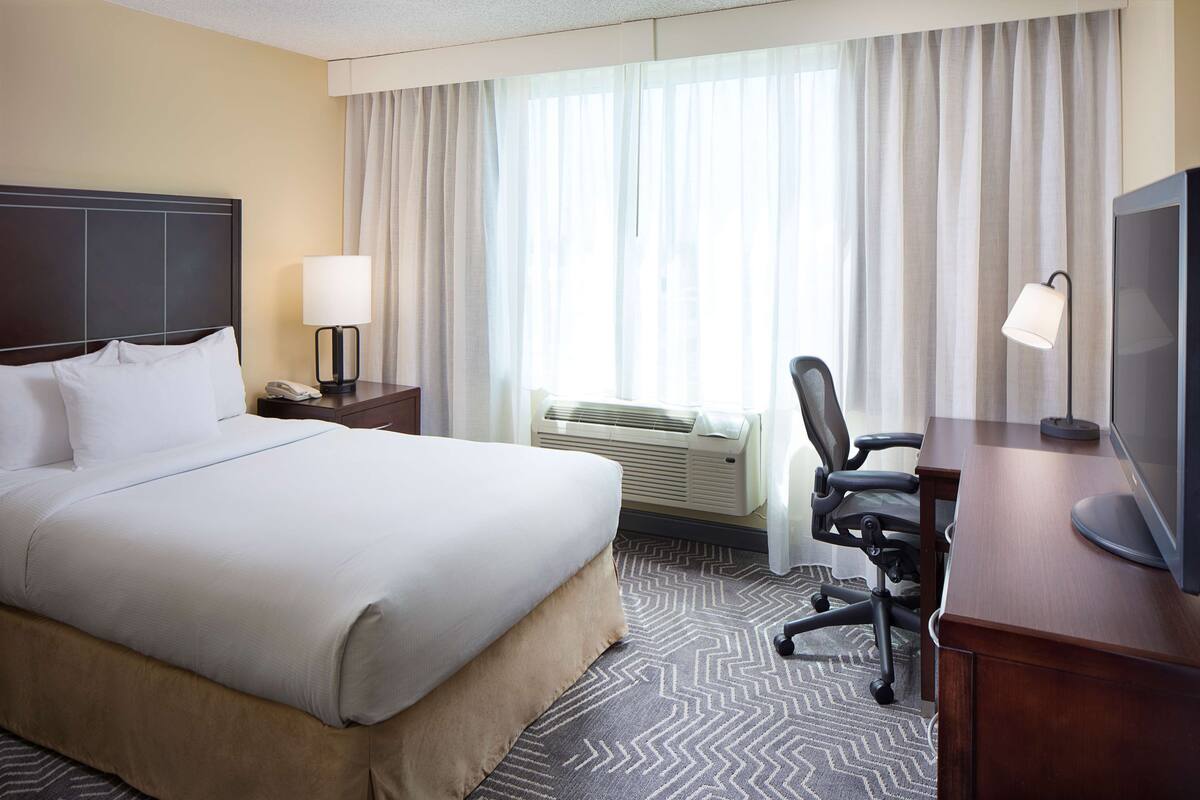 A hotel room with a large bed, a desk with a chair, a television, and a window with curtains.