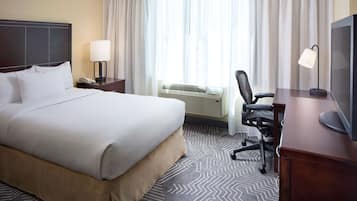 Ein Hotelzimmer mit einem großen Bett, einem Schreibtisch mit Stuhl, einem Fernseher und einem Fenster mit Vorhängen.