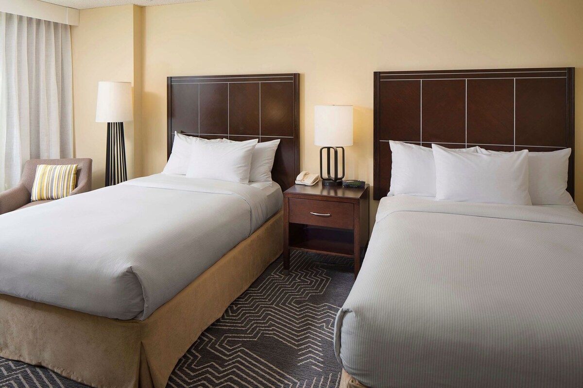 A hotel room with two beds, a nightstand with a lamp, a chair, and a patterned rug.