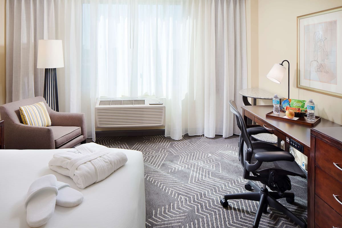A hotel room with a bed, a desk, a chair, an air conditioner, and a lamp.