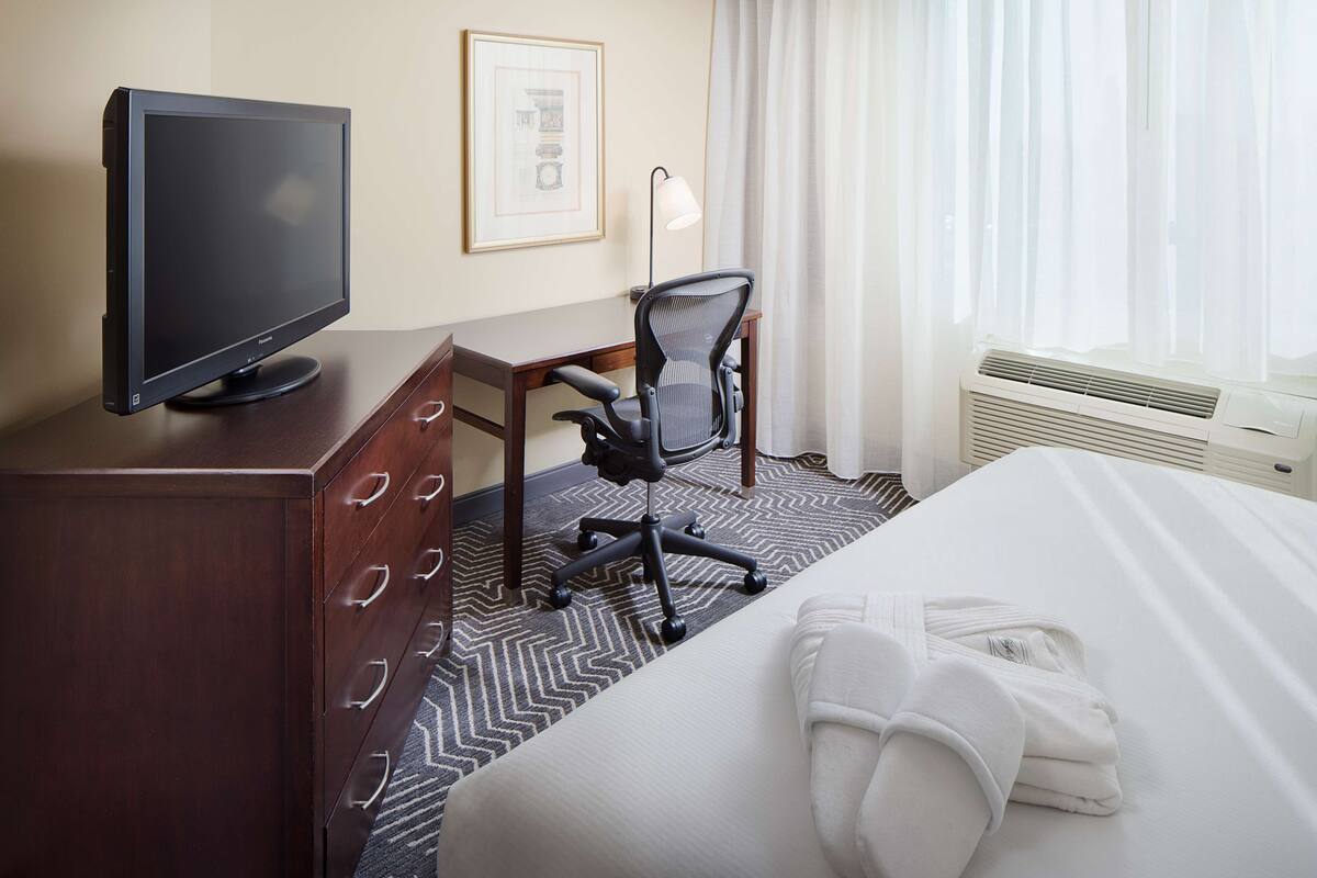 A hotel room with a bed, a desk with a computer monitor, a chair, a desk lamp, and a framed picture on the wall.